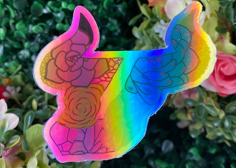 May include: A holographic sticker with a rainbow background featuring a floral design of succulents. The sticker is outlined in black and has a glossy finish.