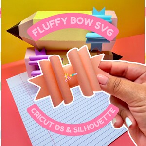 May include: A pink and orange fluffy bow with a pink banner that says "CRICUT DS & SILHOUETTE" on a blue and white lined paper background. The bow is being held by a hand. The image also includes a yellow pencil with a pink banner that says "FLUFFY BOW SVG".