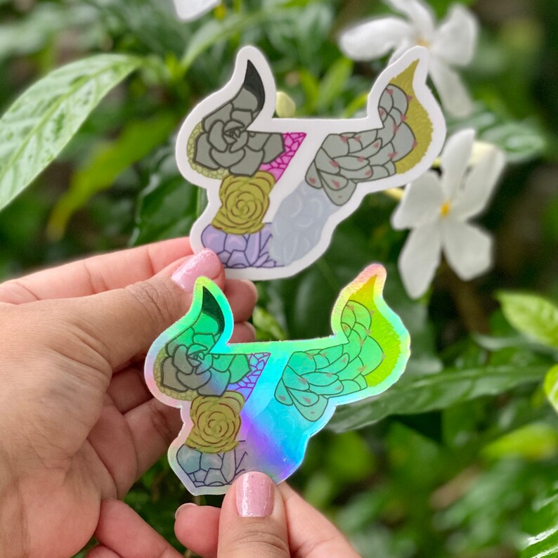 May include: Two stickers of a bull's head made of succulents. One sticker is white with a pink, green, and yellow color scheme. The other sticker is iridescent with a green and yellow color scheme.
