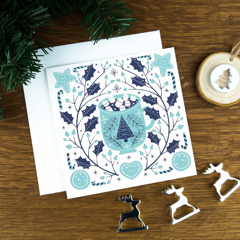 A Christmas card with a mug of hot chocolate in a Scandinavian style illustration. The card sits on an envelope on a wooden background with Christmas trees in the background and three silver reindeer ornaments in the foreground.