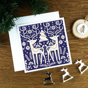 A Christmas card with Scandinavian style illustrations of two reindeers in a Nordic art style. The card sits on an envelope on a wooden background with Christmas trees in the background and three silver reindeer ornaments in the foreground.