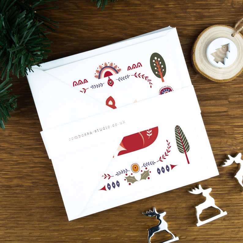 A folk art style Nordic Christmas card pack with festive illustrations surrounding it. The cards sit on a matching white envelope on a wooden table surrounded by little Christmas decorations.