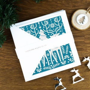 A pack of Christmas cards with  Scandinavian style illustrations. The cards sit on an envelope on a wooden background with Christmas trees in the background and three silver reindeer ornaments in the foreground.