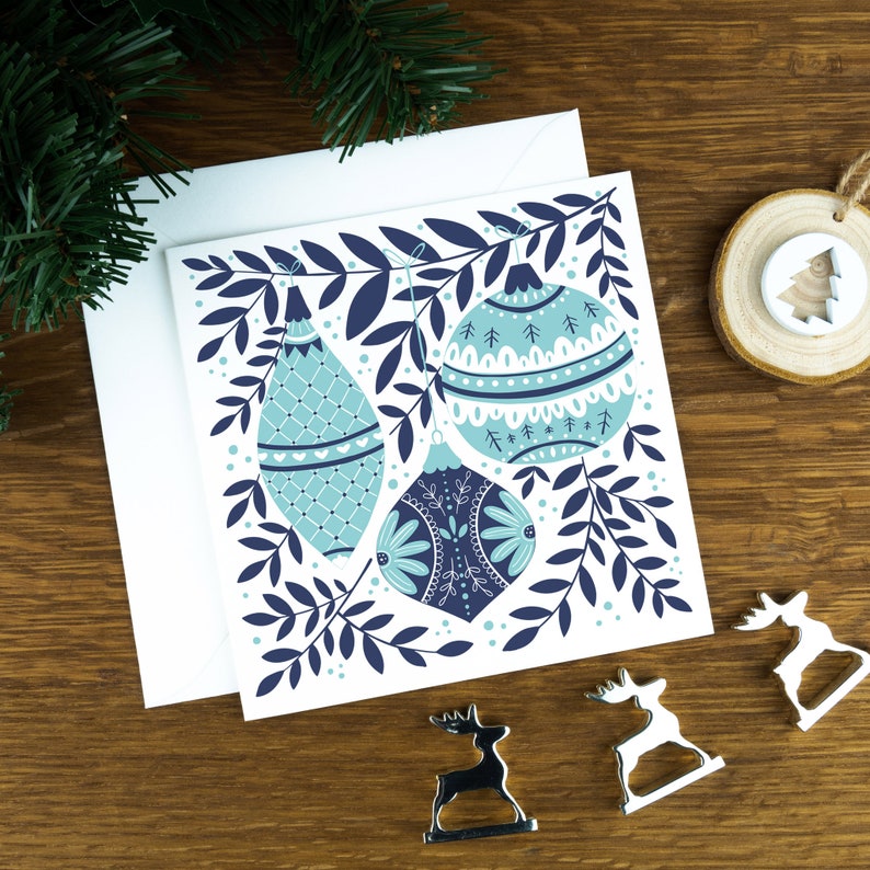 A Christmas card with Christmas decorations in a Scandinavian style illustration. The card sits on an envelope on a wooden background with Christmas trees in the background and three silver reindeer ornaments in the foreground.