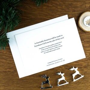 The back of the Christmas card with text on it explaining that a donation will be made to Fareshare UK from the sale of every Christmas card. The card sits on a dark wooden background with Christmas decorations around it.