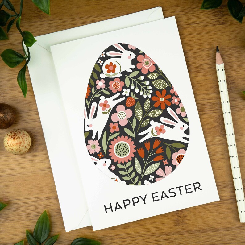 An easter card with a nordic design of an egg filled with a pattern of rabbits and spring flowers. The card says Happy Easter underneath the egg shape. It sits on a wooden board next to two small Easter eggs.