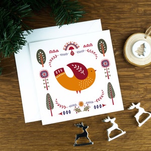 A folk art style Nordic Christmas card with a mustard dove in the middle of it and festive illustrations surrounding it. The card sits on a matching white envelope on a wooden table surrounded by little Christmas decorations.