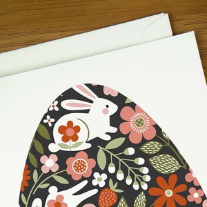 An easter card with a nordic design of an egg filled with a pattern of rabbits and spring flowers. The card says Happy Easter underneath the egg shape. It sits on a wooden board next to two small Easter eggs.