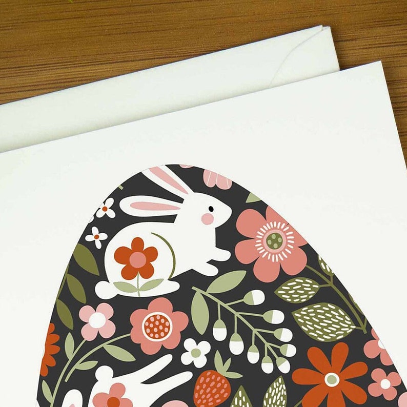 An easter card with a nordic design of an egg filled with a pattern of rabbits and spring flowers. The card says Happy Easter underneath the egg shape. It sits on a wooden board next to two small Easter eggs.