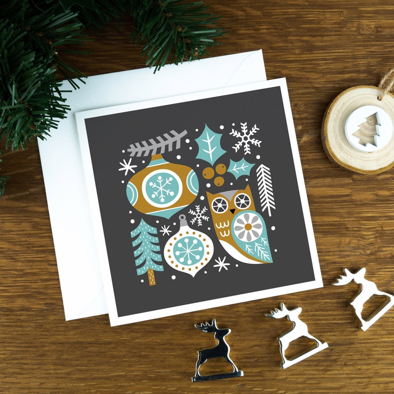 A Christmas card with Scandinavian style illustrations of Christmas decorations and an owl. The card sits on an envelope on a wooden background with Christmas trees in the background and three silver reindeer ornaments in the foreground.