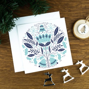 A Christmas card with bells in a Scandinavian style illustration. The card sits on an envelope on a wooden background with Christmas trees in the background and three silver reindeer ornaments in the foreground.