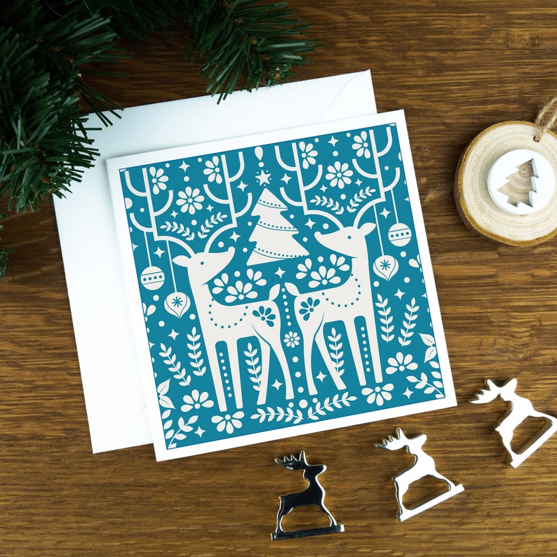 A teal Christmas card with Scandinavian style illustrations of reindeers. The card sits on an envelope on a wooden background with Christmas trees in the background and three silver reindeer ornaments in the foreground.