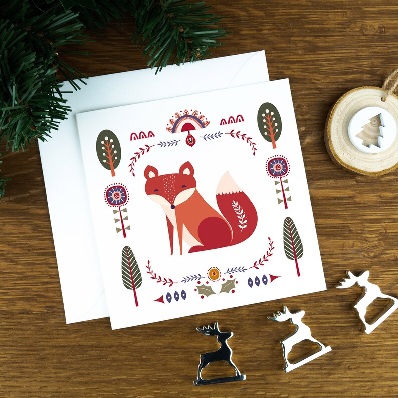 A folk art style Nordic Christmas card with a cute fox in the middle of it and festive illustrations surrounding it. The card sits on a matching white envelope on a wooden table surrounded by little Christmas decorations.