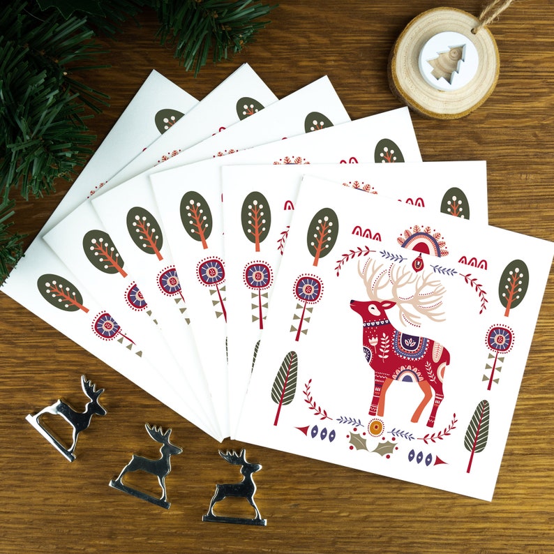 A nordic folk art Christmas card pack with a red deer illustration in the centre and Christmas trees and decorations surrounding it. The cards sit on matching envelopes on a wooden background.