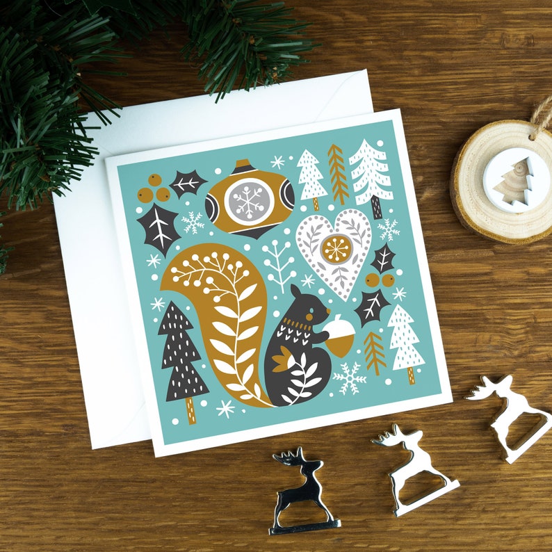 A Christmas card in a nordic style featuring a squirrel and a variety of festive decorations. The card sits on a matching envelope on a wooden background with Christmas decorations surrounding it. The background of the card is teal.