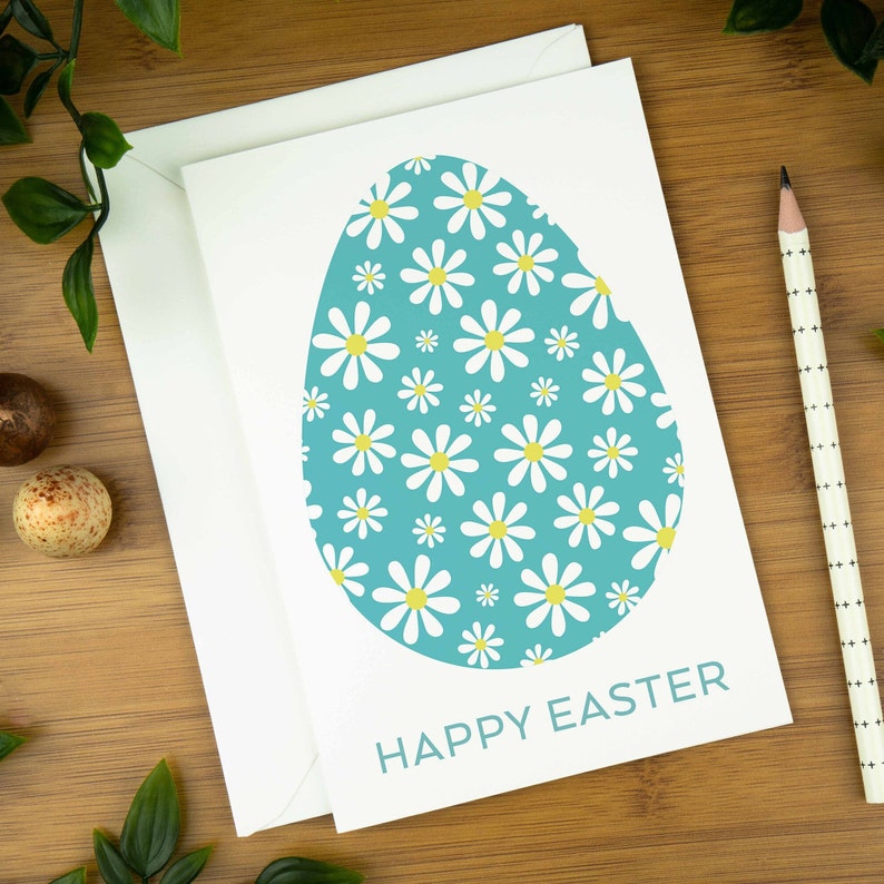 An easter card with a nordic design of an egg filled with a spring daisy pattern. The card says Happy Easter underneath the egg shape. It sits on a wooden board next to two small Easter eggs.