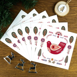 A folk art Nordic Christmas card pack with a red dove of peace in the centre. The cards sit on matching white envelopes on a wooden background with Christmas decorations surrounding it.