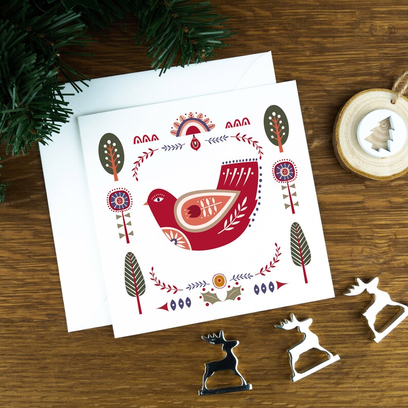 A folk art style Nordic Christmas card with a red dove in the middle of it and festive illustrations surrounding it. The card sits on a matching white envelope on a wooden table surrounded by little Christmas decorations.