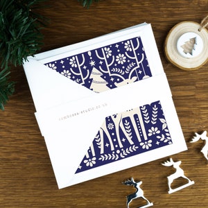 A pack of Christmas cards with  Scandinavian style illustrations. The cards sit on an envelope on a wooden background with Christmas trees in the background and three silver reindeer ornaments in the foreground.
