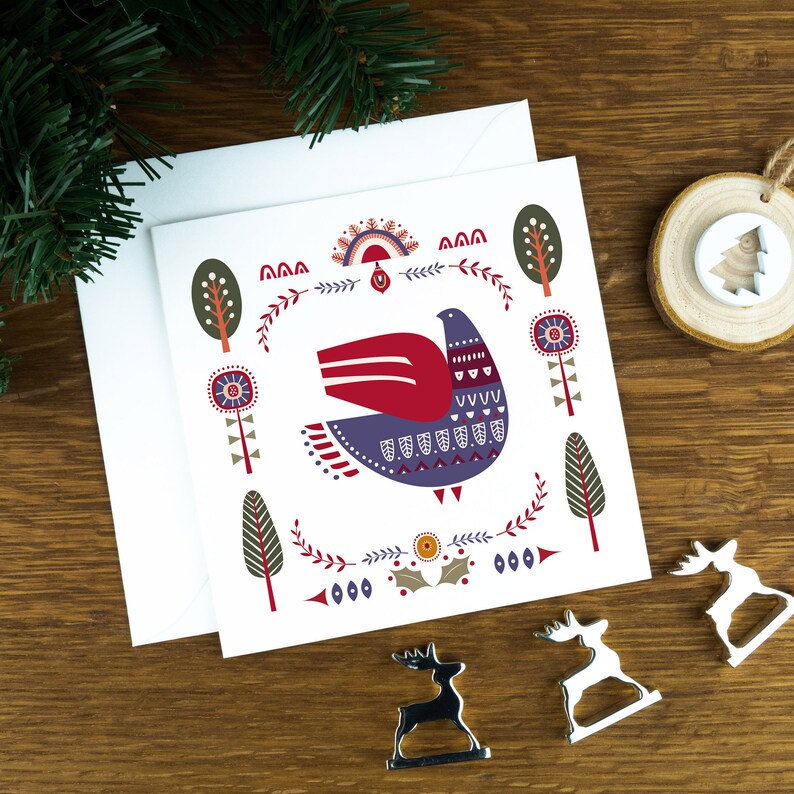 A folk art style Nordic Christmas card with a purple dove in the middle of it and festive illustrations surrounding it. The card sits on a matching white envelope on a wooden table surrounded by little Christmas decorations.