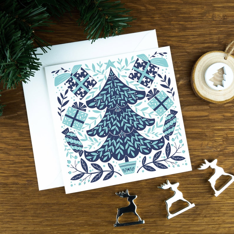 A Christmas card with a Christmas tree in a Scandinavian style illustration. The card sits on an envelope on a wooden background with Christmas trees in the background and three silver reindeer ornaments in the foreground.