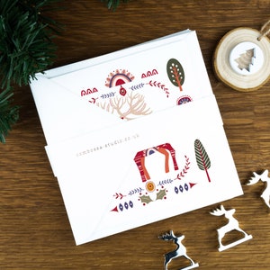 A nordic folk art Christmas card pack with a red deer illustration in the centre and Christmas trees and decorations surrounding it. The cards sit on matching envelopes on a wooden background.