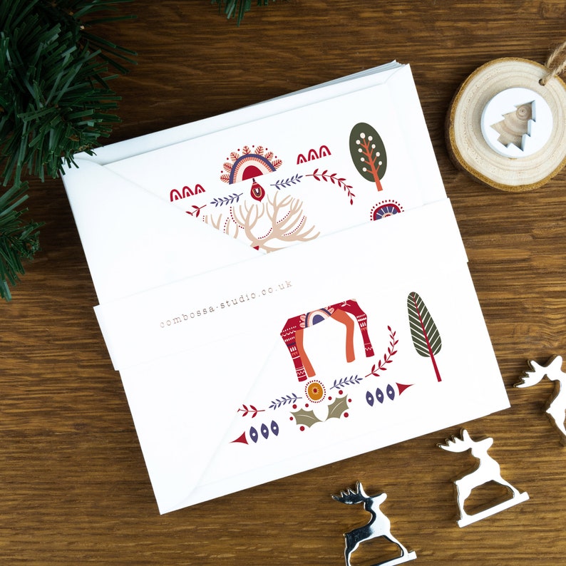 A nordic folk art Christmas card pack with a red deer illustration in the centre and Christmas trees and decorations surrounding it. The cards sit on matching envelopes on a wooden background.