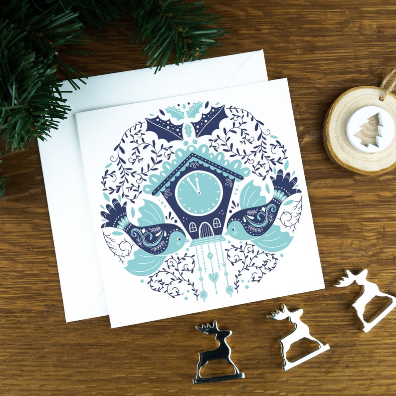 A Christmas card with birds and a clock in a Scandinavian style illustration. The card sits on an envelope on a wooden background with Christmas trees in the background and three silver reindeer ornaments in the foreground.