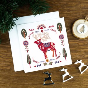 A folk art style Nordic Christmas card with a reindeer in the middle of it and festive illustrations surrounding it. The card sits on a matching white envelope on a wooden table surrounded by little Christmas decorations.