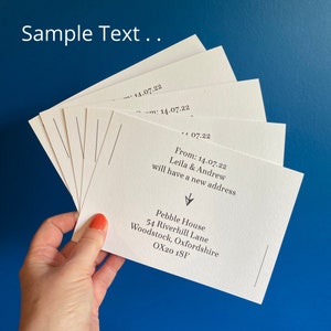 The reverse of the change of address cards are being held in a fan shape against a blue wall. The cards have sample text on them - "From 14.07.22 Leila and Andrew will have a new address." The rest of the text specifies their new address.