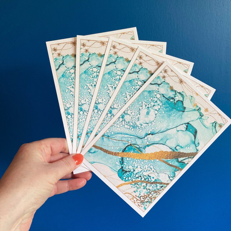 A hand holding a display of five of the change of address cards. The front of the card is displayed and has an abstract art blue and gold design on it. The cards are being held up against a blue wall.