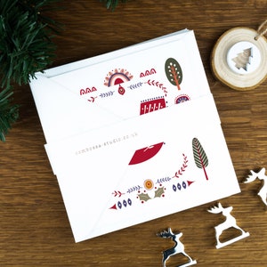 A folk art Nordic Christmas card pack with a red dove of peace in the centre. The cards sit on matching white envelopes on a wooden background with Christmas decorations surrounding it.