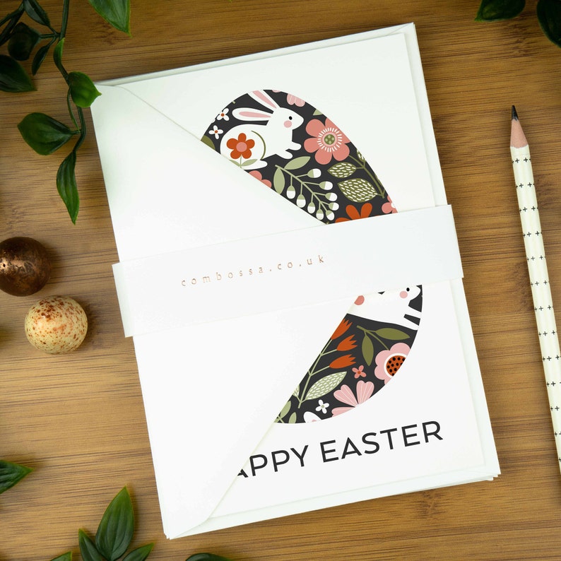 An easter card with a nordic design of an egg filled with a pattern of rabbits and spring flowers. The card says Happy Easter underneath the egg shape. It sits on a wooden board next to two small Easter eggs.