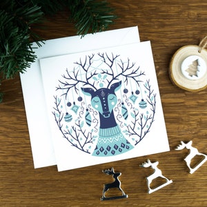A Christmas card with a reindeer in a Scandinavian style illustration. The card sits on an envelope on a wooden background with Christmas trees in the background and three silver reindeer ornaments in the foreground.