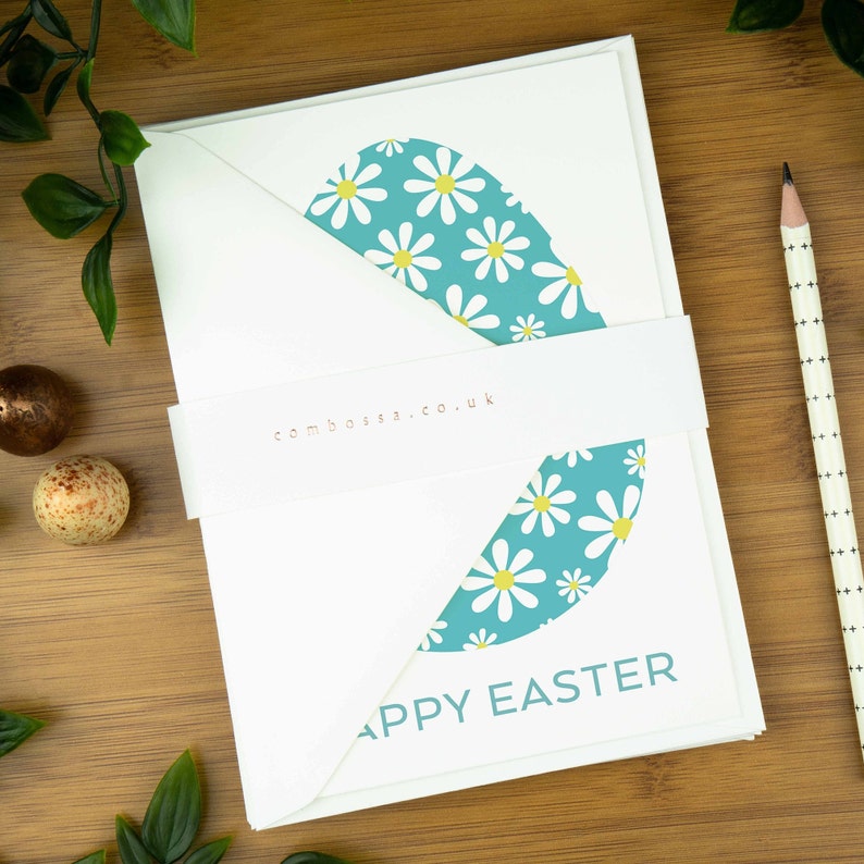 An easter card with a nordic design of an egg filled with a spring daisy pattern. The card says Happy Easter underneath the egg shape. It sits on a wooden board next to two small Easter eggs.