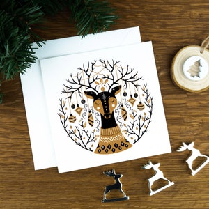 A Christmas card with Scandinavian illustrations of a reindeer in a Nordic art style. The card sits on an envelope on a wooden background with Christmas trees in the background and three silver reindeer ornaments in the foreground.
