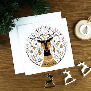 A Christmas card with Scandinavian illustrations of a reindeer in a Nordic art style. The card sits on an envelope on a wooden background with Christmas trees in the background and three silver reindeer ornaments in the foreground.