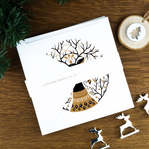 A pack of Christmas cards with  Scandinavian style illustrations. The cards sit on an envelope on a wooden background with Christmas trees in the background and three silver reindeer ornaments in the foreground.