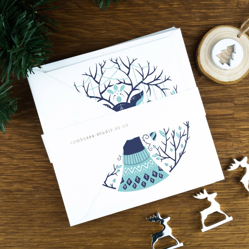 A pack of Christmas cards with  Scandinavian style illustrations. The cards sits on an envelope on a wooden background with Christmas trees in the background and three silver reindeer ornaments in the foreground.