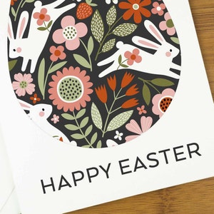 An easter card with a nordic design of an egg filled with a pattern of rabbits and spring flowers. The card says Happy Easter underneath the egg shape. It sits on a wooden board next to two small Easter eggs.