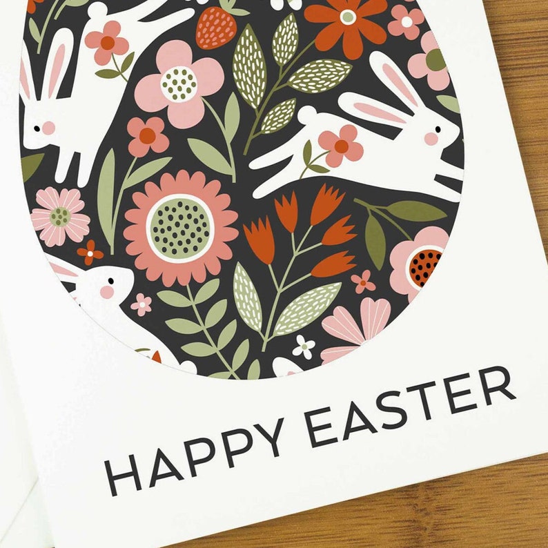 An easter card with a nordic design of an egg filled with a pattern of rabbits and spring flowers. The card says Happy Easter underneath the egg shape. It sits on a wooden board next to two small Easter eggs.