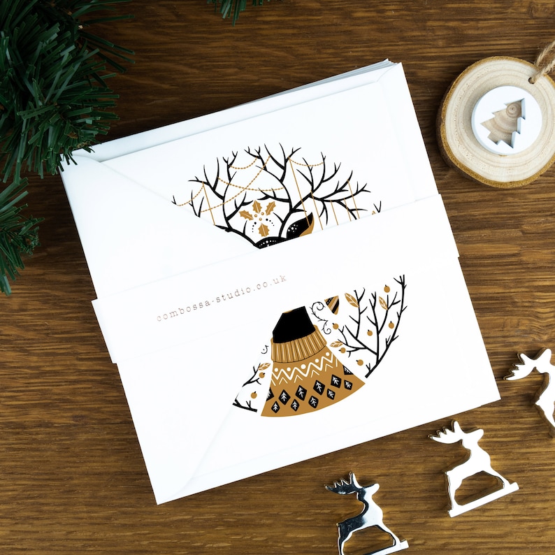 A pack of Christmas cards with  Scandinavian style illustrations. The cards sit on an envelope on a wooden background with Christmas trees in the background and three silver reindeer ornaments in the foreground.