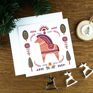 A folk art style Nordic Christmas card with an illustrated animal in the middle of it and festive illustrations surrounding it. The card sits on a matching white envelope on a wooden table surrounded by little Christmas decorations.