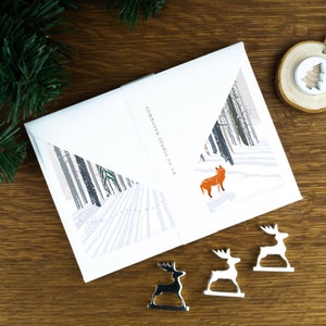 A Christmas card with a fox looking into trees in a snowy landscape illustration. The card sits on an envelope on a wooden background with Christmas trees in the background and three silver reindeer ornaments in the foreground.