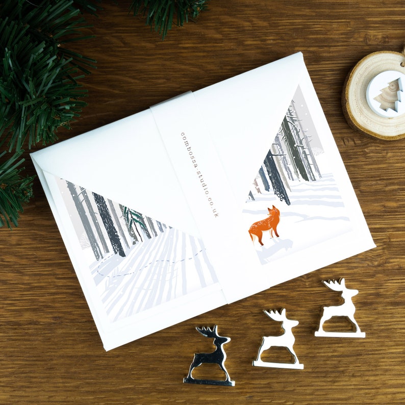 A Christmas card with a fox looking into trees in a snowy landscape illustration. The card sits on an envelope on a wooden background with Christmas trees in the background and three silver reindeer ornaments in the foreground.
