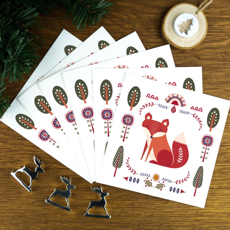 A folk art Nordic Christmas card pack with festive illustrations. The cards sit on a matching white envelope on a wooden table surrounded by little Christmas decorations. The top card has a fox on it, each of the six designs are different.