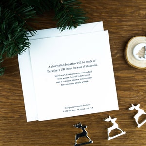 The back of a Christmas card with the message about the donation to FareShare UK. The card sits on an envelope on a wooden background with Christmas trees in the background and three silver reindeer ornaments in the foreground.