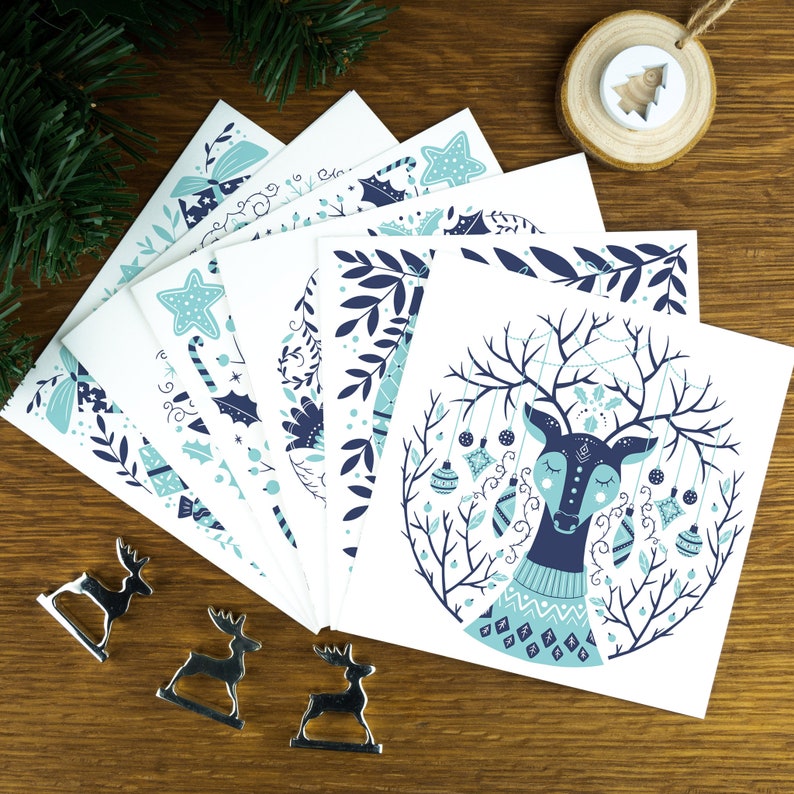 A pack of Christmas cards with  Scandinavian style illustrations. The cards sits on an envelope on a wooden background with Christmas trees in the background and three silver reindeer ornaments in the foreground.