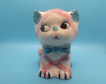Puede incluir: Una figurita de cerámica de un gato con rayas rosas y azules, un lazo azul y ojos negros. El gato está sentado sobre un fondo azul.