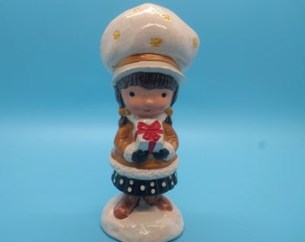 May include: A ceramic figurine of a girl wearing a white hat with yellow flowers, a brown coat, and a black and white polka dot skirt. She is holding a red and white gift box with a red bow. The figurine is standing on a white base.